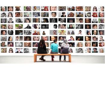 Photo of three people sitting on a bench with their backs to the viewer, looking at a gallery wall of photographs of different faces. The same as the Personality Psychology image.